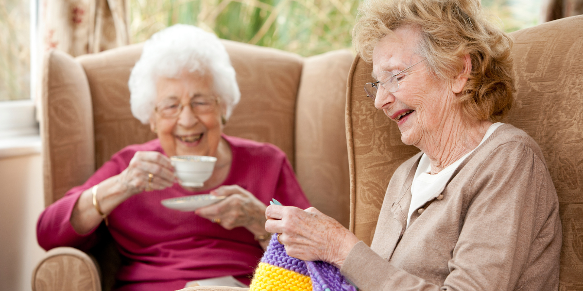 dos-personas-mayores-sonriendo-en-una-residenca Dos personas adultas mayores sonriendo, una de ellas teje, mientras la otra toma un té