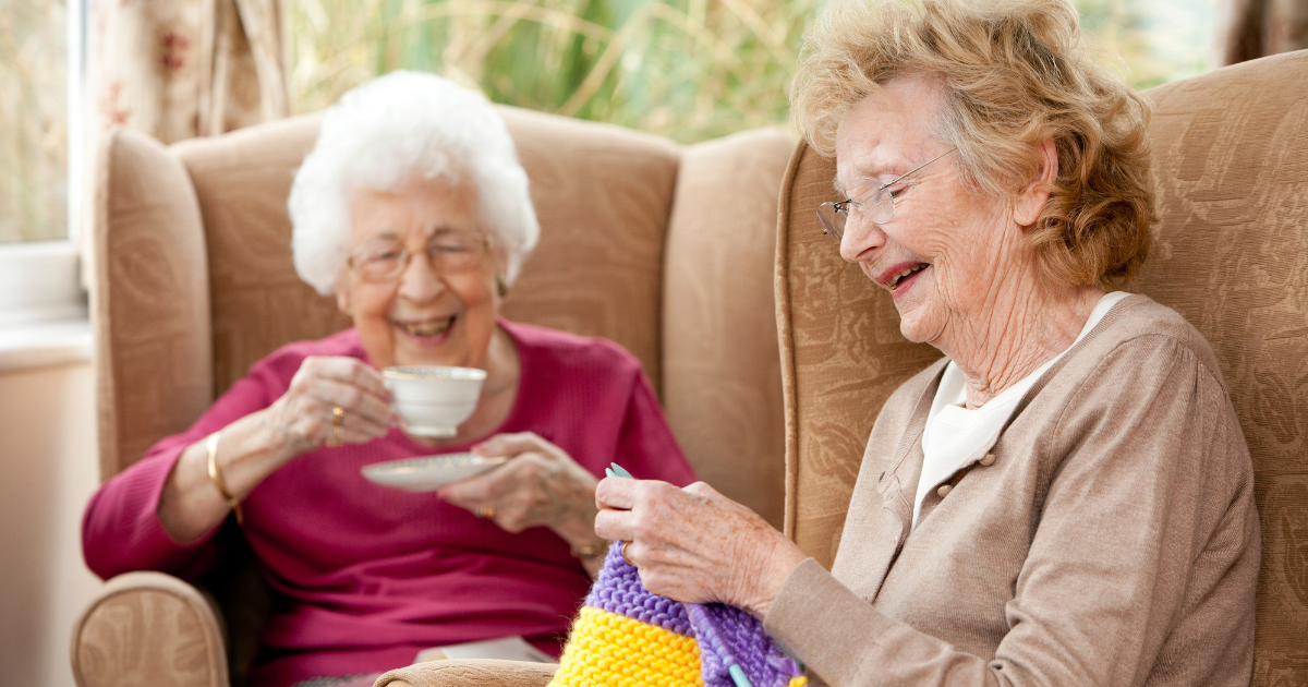 dos-personas-mayores-sonriendo-en-una-residenca Dos personas adultas mayores sonriendo, una de ellas teje, mientras la otra toma un té